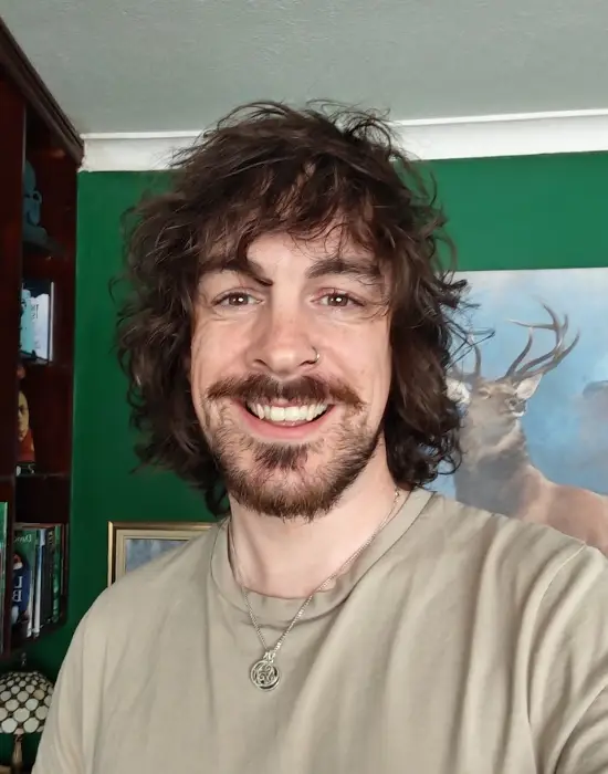 Non Officer Director, Clinton Burn, is a white skinned man with medium length loose curled chestnut coloured hair and brown eyes. He is smiling and standing before a bookshelf adjacent to a green wall adorned with artwork of a stag on a blue background.