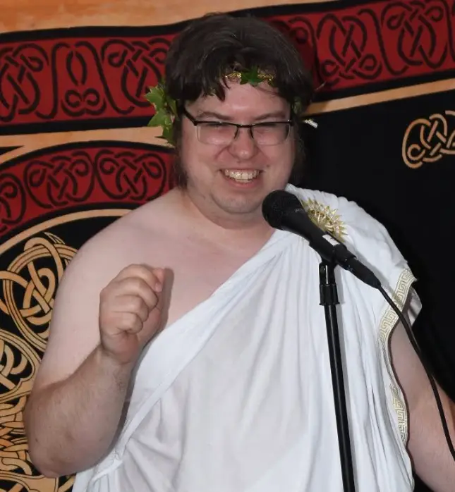 Chief, Council of Regional Druids, Dave "Thexalon" Kleinschmidt is a white skinned man with dark hair and wears rimless glasses. He standing in front of a red, black and beige tapestry inlaid with knotwork designs, is smiling and wearing a white toga and a leaf crown.