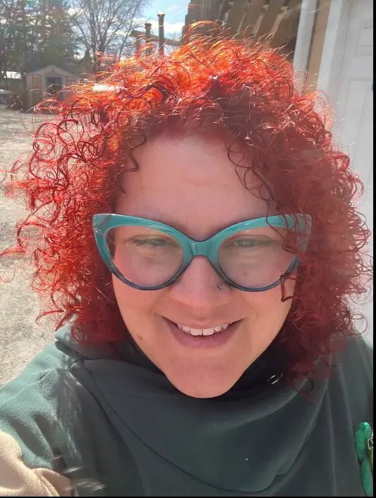 Chief, Council of Senior Druids, Tasha Smith is smiling. She is a white skinned woman with bright curly hair, wearing a forest green cowl-necked sweater and teal cat-eye glasses.