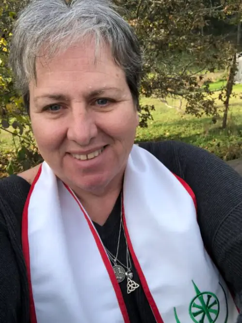 Vice Archdruid, Rev. Diane Cacciato us a white skinned woman who is smiling, wearing a white stole embroidered with a green sigil.
