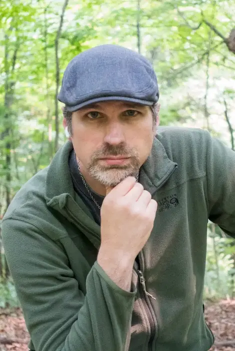 Non-Officer Director, Rev. Michael J Dangler is a white skinned man sporting a light brown goatee, wearing a blue paperboy hat and a green sweater. He is leaning forward with his chin perched on his index and thumb in a well lit forested area.