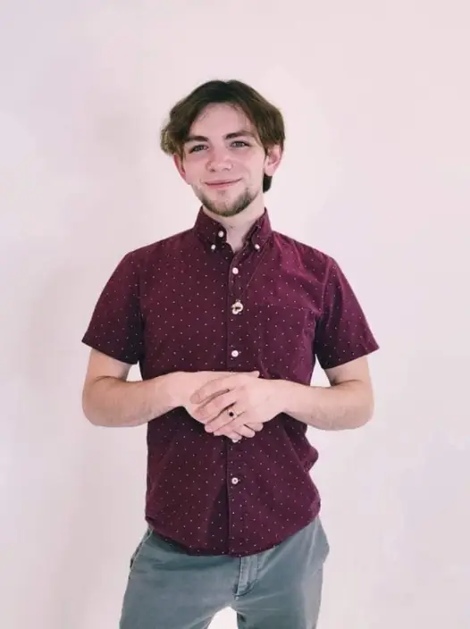 Member's Advocate, Torre Journey, is a white skinned man with short dark hair and a dark goatee. he is smiling, wearing a burgundy shirt and standing before a light pink background holding his hands before him above his waist.